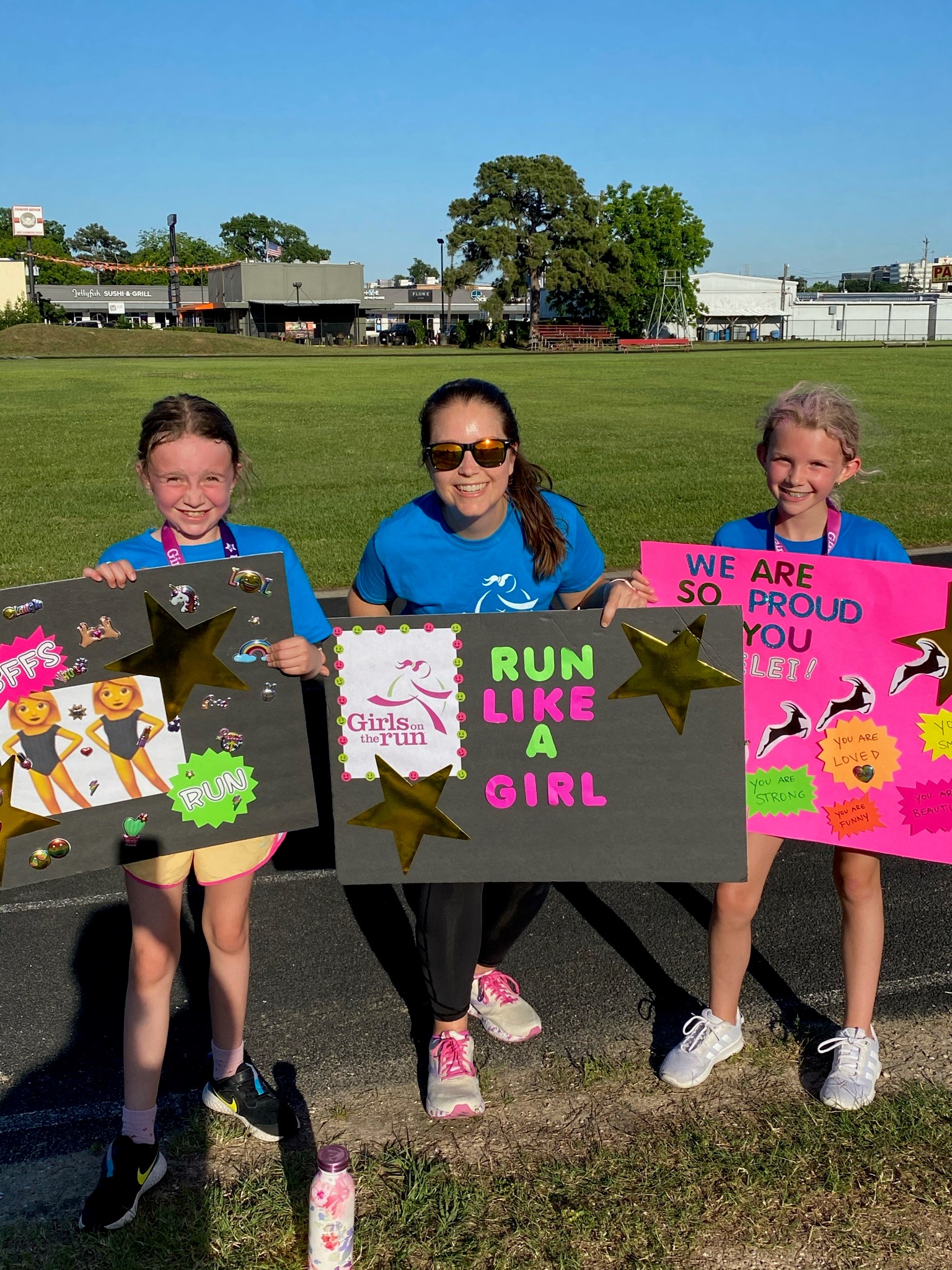 Girls on the Run Coach and participants standing outdoors, each holding a handmade sign. The signs have power poses, positive affirmation statements and words of support.