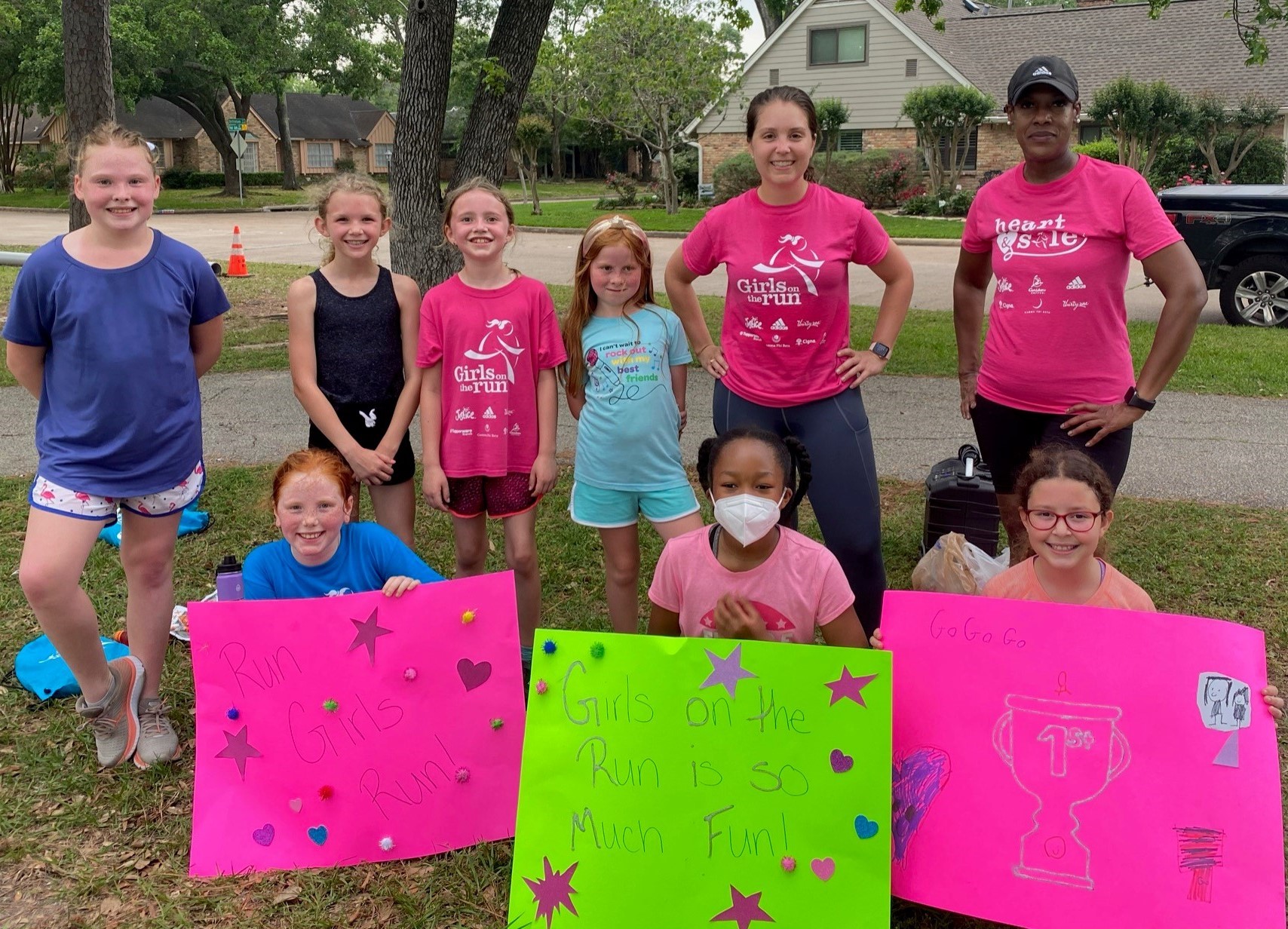 Girls on the Run coaches and teammates grouped together holding signs with positive affirmations about GOTR. They read: "Girls on the Run is so much fun!" and "Go, go, go!" Great reminders that no matter our pathway, we can support each other.