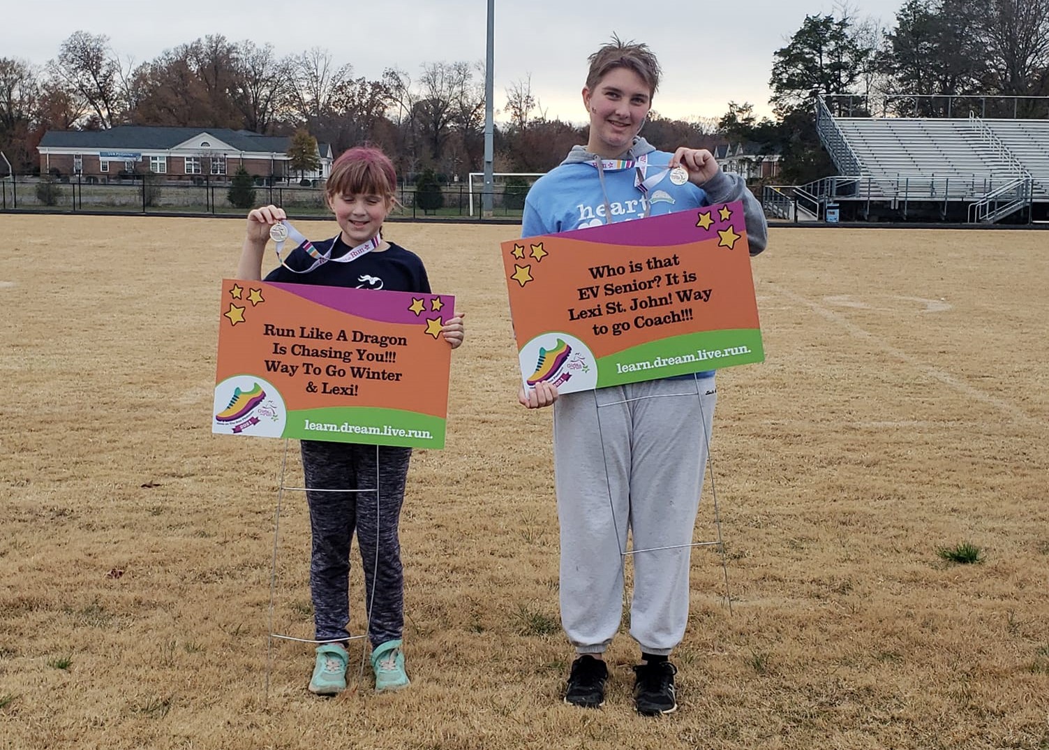 Participants holding up a sign and coach holding up a fantastic 5K sign on a football field. Both are holding metals too.