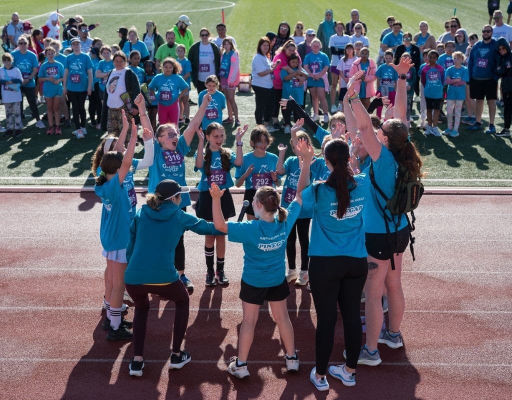 A Girls on the Run team in Southwest Missouri gathers in a joyful cheer before their 5K, surrounded by supportive families and community members. The energy is high, the smiles are wide, and the celebration of confidence and teamwork is in full swing!