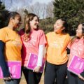 Four Girls on the Run coaches standing outdoors, smiling and laughing together while holding program curriculum guides.