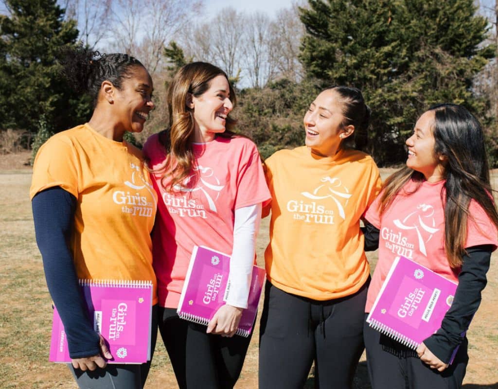 Four Girls on the Run coaches standing outdoors, smiling and laughing together while holding program curriculum guides.