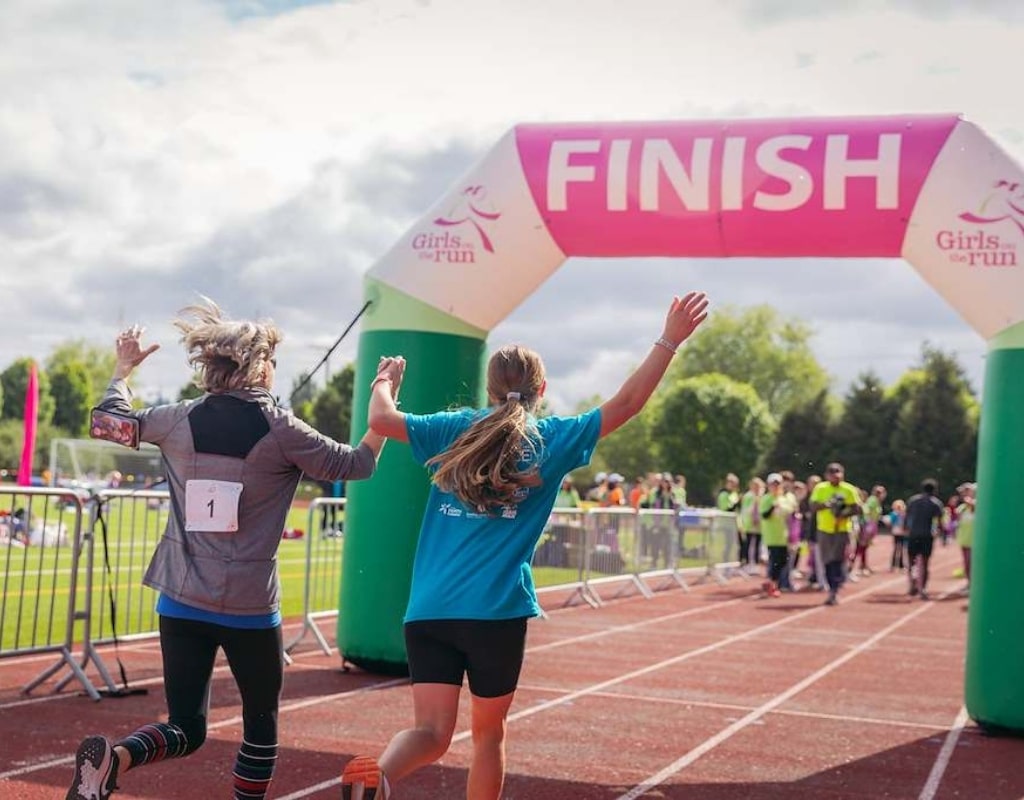 Mother and daughter cross the finish line at the Girls on the Run 5K hand in hand, with their arms raised above their heads as they joyfully cross the finish line together.