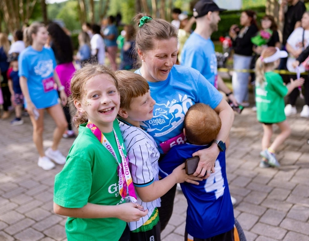 Girl wearing a green Girls on the Run shirt smiles at the camera as she hugs her mother and brothers at the end of the Girls on the Run 5K. She is wearing a medal with a big smile on her face.