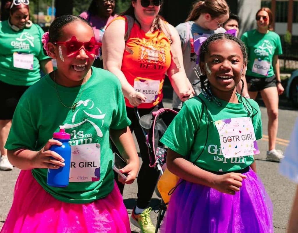 Two girls wearing green Girls on the Run shirts and purple tutus smile as they run outside with a group during the 5K.
